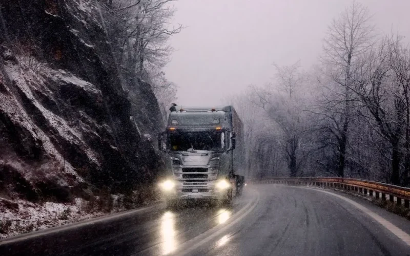 Heavy truck driving through snowy mountain road during winter snowfall.