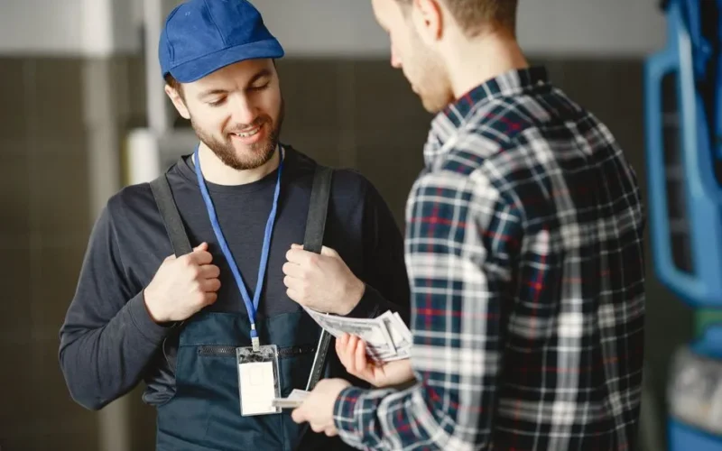 A mechanic in a blue cap receives cash payment from a customer in a plaid shirt, smiling.