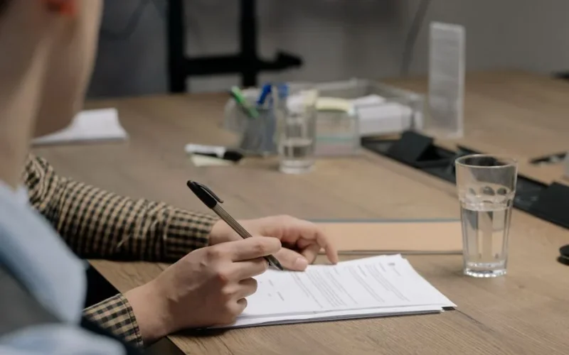 Person in business attire signing a document at a wooden table in an office setting.