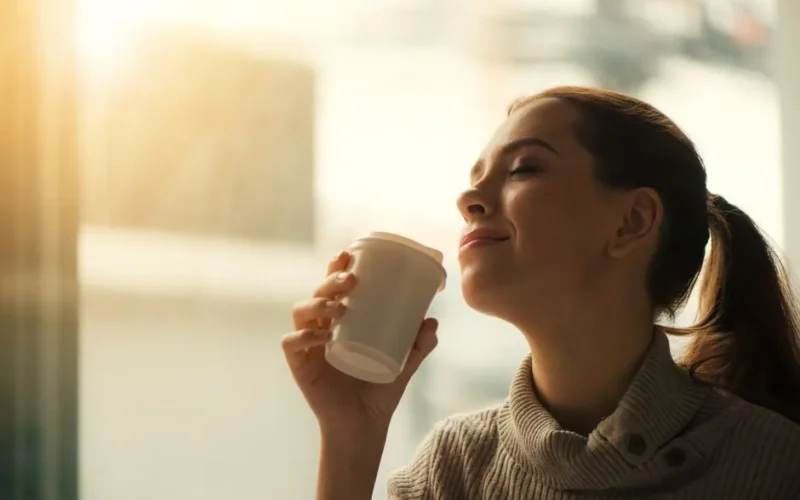 Smiling woman savoring morning coffee by the window in soft warm light.