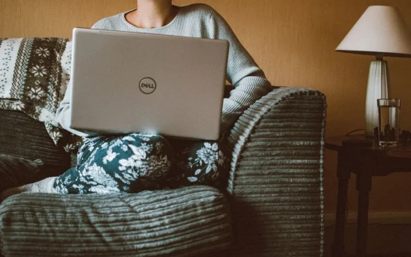A woman in casual clothing works on a laptop while relaxing on a cozy sofa indoors.