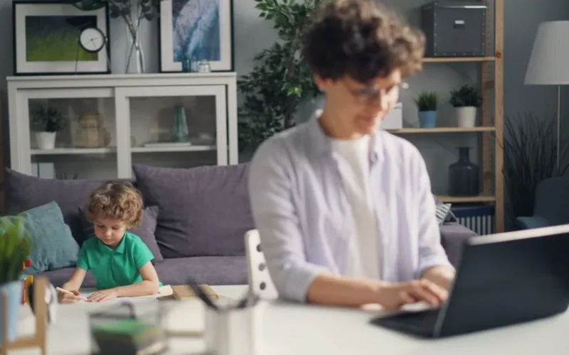 Mother Working on Laptop and Son Playing behind