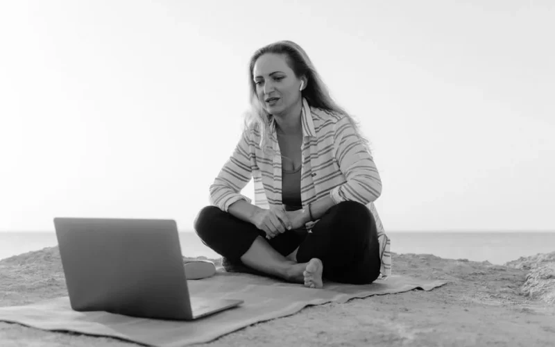 A woman sits on a beach, using a laptop for a video call, offering a peaceful working environment.