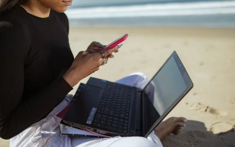 Person multitasking with a laptop and smartphone by the beach, embracing remote work lifestyle.