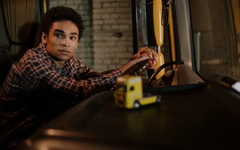 A young man sits in the cabin of a truck, hands on the steering wheel, with a model truck on the dashboard.