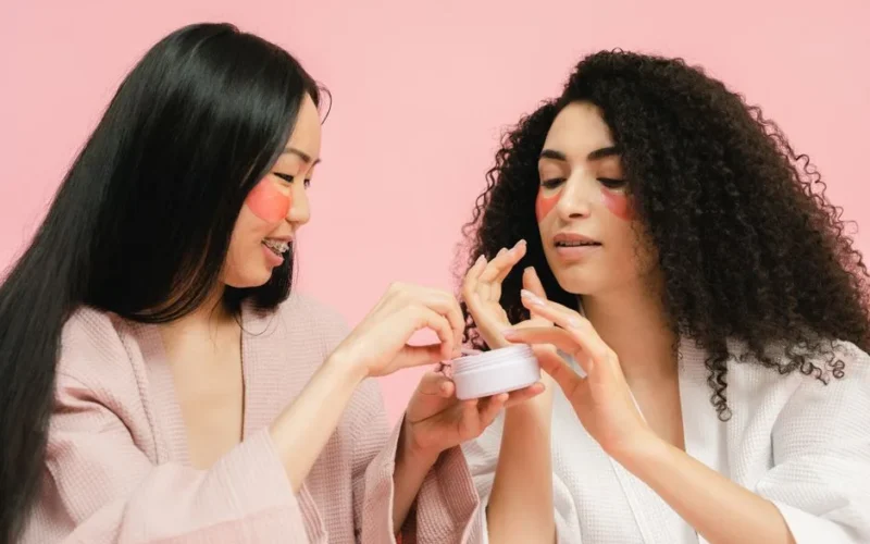 Two women with facial patches apply skincare cream, enhancing beauty and self-care routine.
