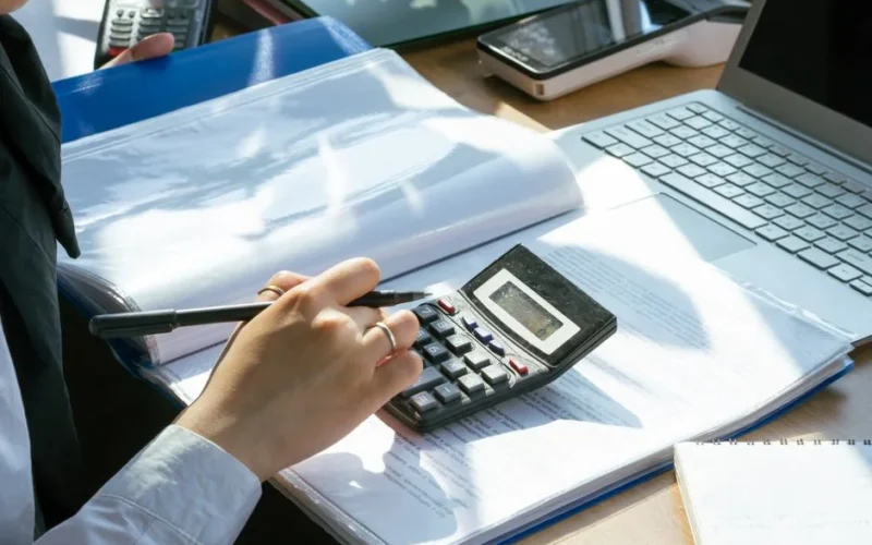 A person working on financial calculations using a calculator and laptop at an office desk.