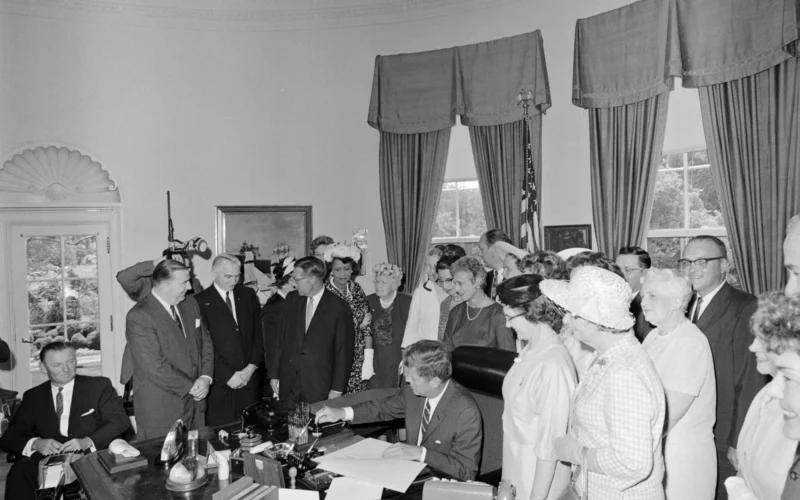 In this photograph President John F. Kennedy signs the Equal Pay Act in the Oval Office of the White House, Washington, D.C. An unidentified stenographer sits at far left. Others (L-R): unidentified stenographer (seated); Senator Jennings Randolph (West Virginia); Senator Wayne Morse (Oregon); Representative Florence Price Dwyer of New Jersey (in back, mostly hidden); Senator Winston L. Prouty (Vermont); President of the National Council of Negro Women (NCNW), Dorothy Height; former Director of the Women’s Bureau in the Department of Labor, Mary Anderson; Assistant Secretary of Labor for Labor Standards, Esther Peterson; Director of Research and Program Resources for the Young Women’s Christian Association (YWCA), Ethlyn Christensen; President of the National Council of Jewish Women (NCJW), Pearl Larner Willen; Representative Carlton Sickles of Maryland (in back); Representative Leonor K. Sullivan of Missouri (in back mostly hidden); President of the National Federation of Business and Professional Women’s Clubs (NFBPWC), Dr. Minnie Miles (in front); Executive Director of the National Council of Catholic Women (NCCW), Margaret Mealey; Representative Robert P. Griffin of Michigan (in back, partially hidden); Representative Julia Butler Hansen (Washington); Representative James G. O’Hara (Michigan); Representative Edith Green of Oregon (mostly hidden); Director of United Automobile Workers (UAW) Women’s Department, Caroline Davis (on edge of frame, foreground); Vice President Lyndon B. Johnson (on edge of frame, behind Ms. Davis).