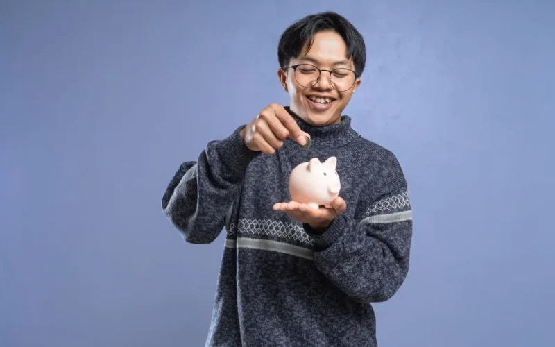 Smiling man holding a piggy bank and inserting a coin, representing savings and financial planning.