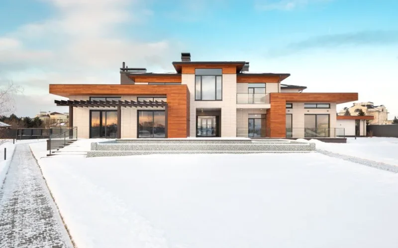 Exterior design of modern cottage house facade with large windows and mixed wooden and stone cladding and spacious yard in snowy winter suburban area