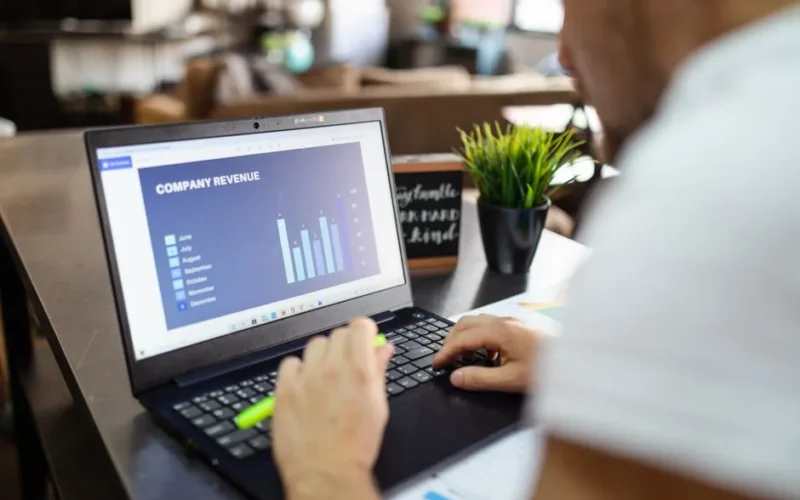 Man using a laptop to analyze company revenue data with graph charts in office setting.