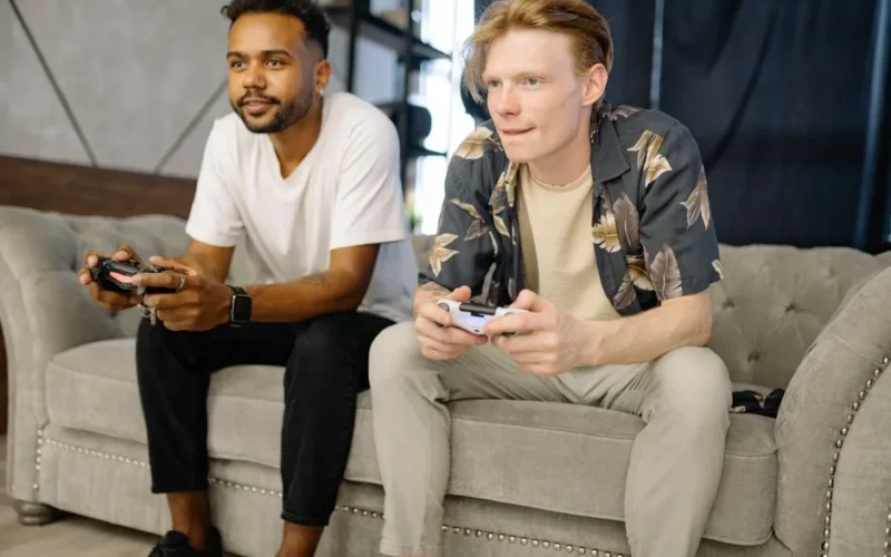 Two friends sitting on a sofa, playing video games together at home.