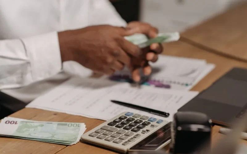 Hands counting euro bills on a wooden desk with calculator, financial documents, and laptop nearby.