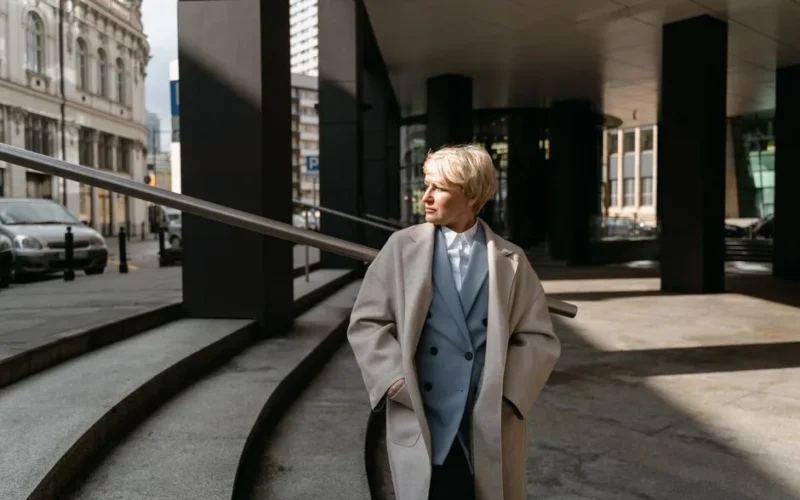 Elegant woman in a trench coat, standing outdoors in a modern urban environment during daytime.
