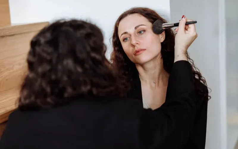 A woman uses a makeup brush to apply cosmetics to another woman in a cozy indoor setting.