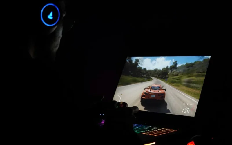 A gamer using a laptop and headphones to play a car racing game in a dark setting.