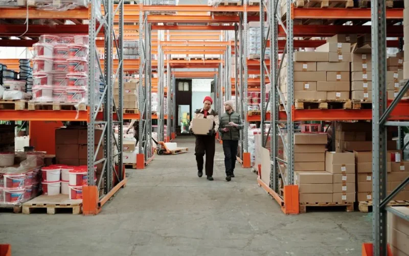 Two workers handle a package in a spacious warehouse surrounded by shelves stocked with boxes and products.