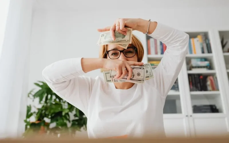 Smiling woman indoors, playfully holding cash in hands, expressing joy.