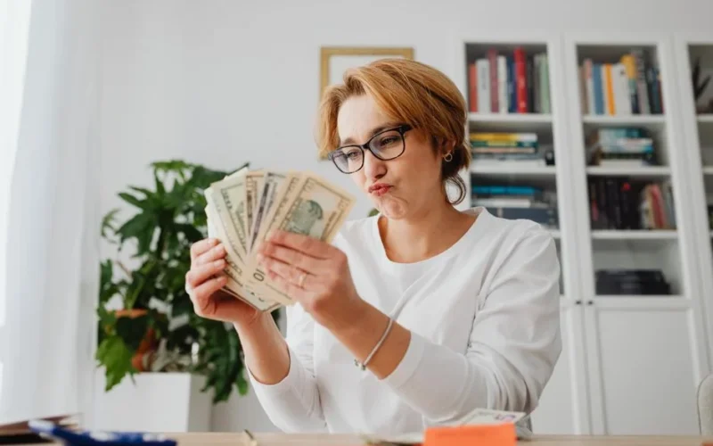 Woman wearing glasses, counting banknotes with a focused expression in a home office setting.