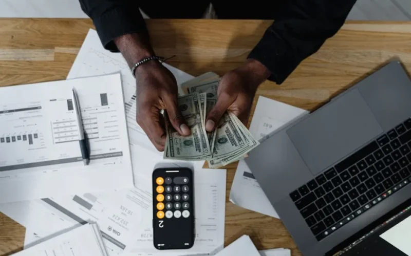 Person counting US dollars, using a calculator and laptop, with financial documents on a wooden desk.