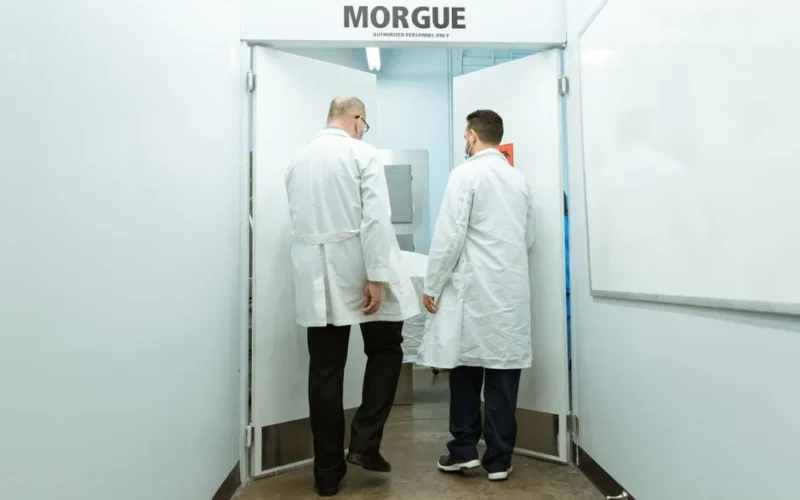 Medical professionals in lab coats entering a morgue with a body on stretcher.