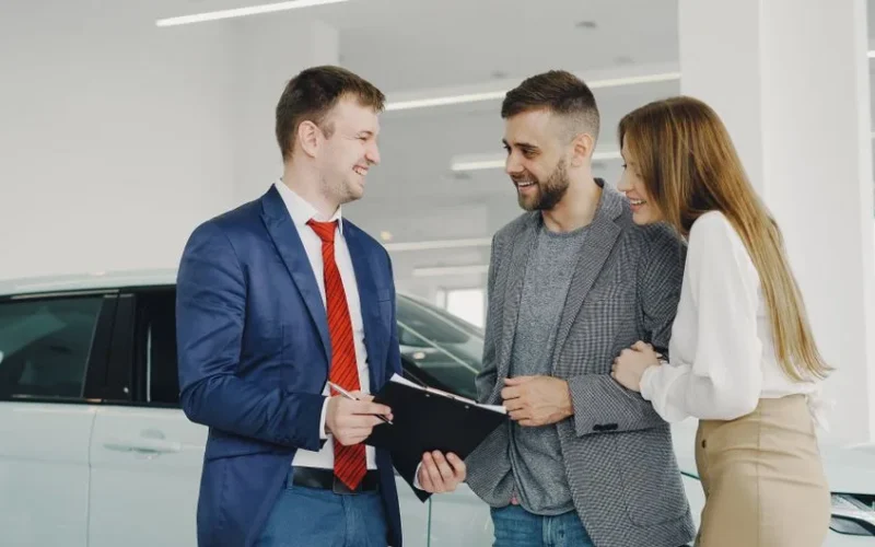 A smiling couple buys a new car from a confident salesman inside a modern car dealership.