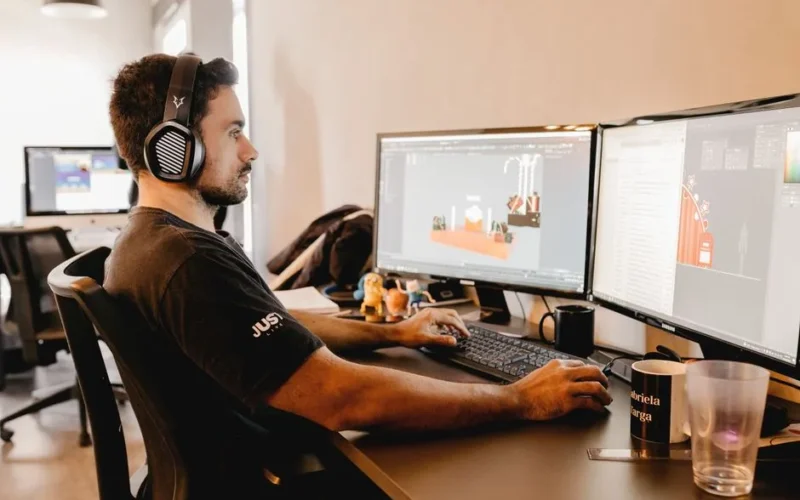 A man wearing headphones works on a computer with two monitors in a modern office setup.