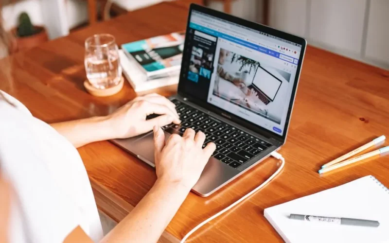 A person working remotely on a laptop at a wooden desk with stationery and a notepad.