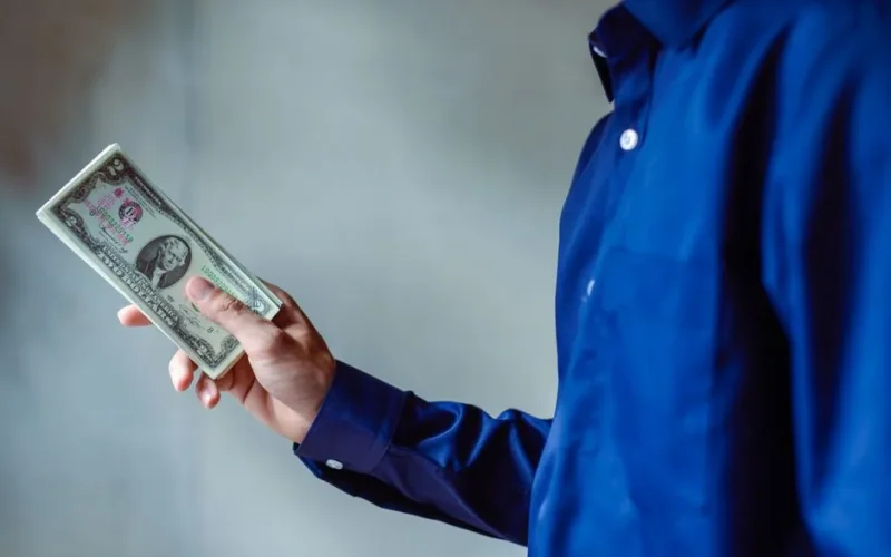 A person in a blue shirt holding a stack of US dollar bills, symbolizing wealth and finance.