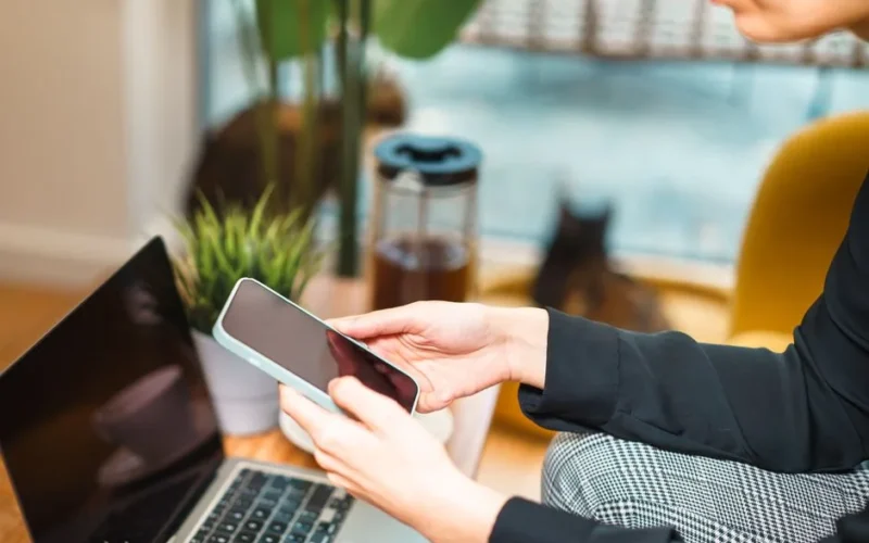 Woman working remotely from home, using a smartphone and laptop at a cozy desk with a plant and cat in background.
