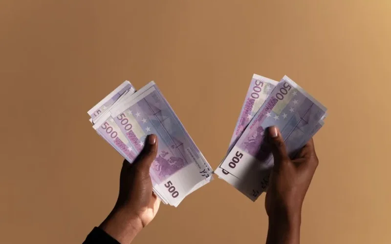 Close-up image of hands holding 500 euro banknotes on a neutral beige background.