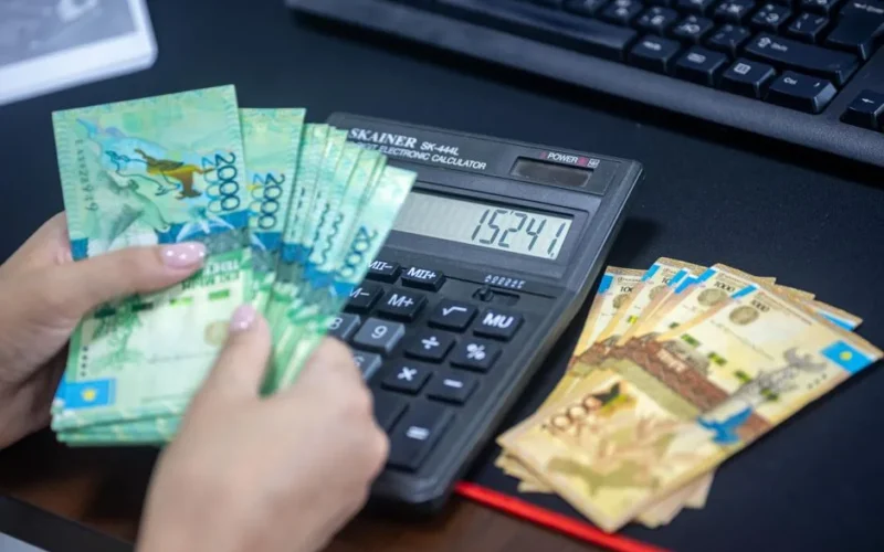 Hands counting Kazakhstani tenge while using calculator at desk.