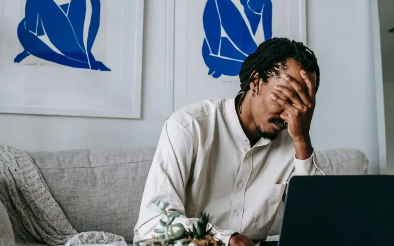 Frustrated young bearded African American male freelancer with dreadlocks in casual shirt working on laptop at home and covering eyes with hand after failure