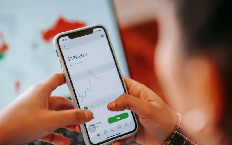 Crop anonymous trader using app on cellphone with graph and dollar price on screen in house