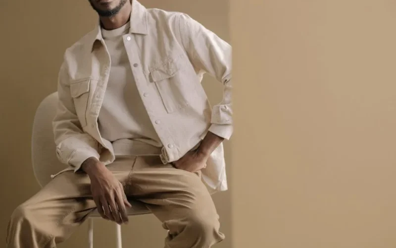 Minimalistic portrait of a man wearing a beige jacket and pants, sitting casually indoors with neutral tones.