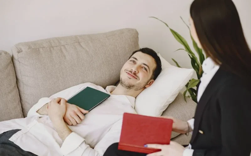 Positive man in formal wear lying on sofa with notebook in hands while smiling and talking with therapist in black suit