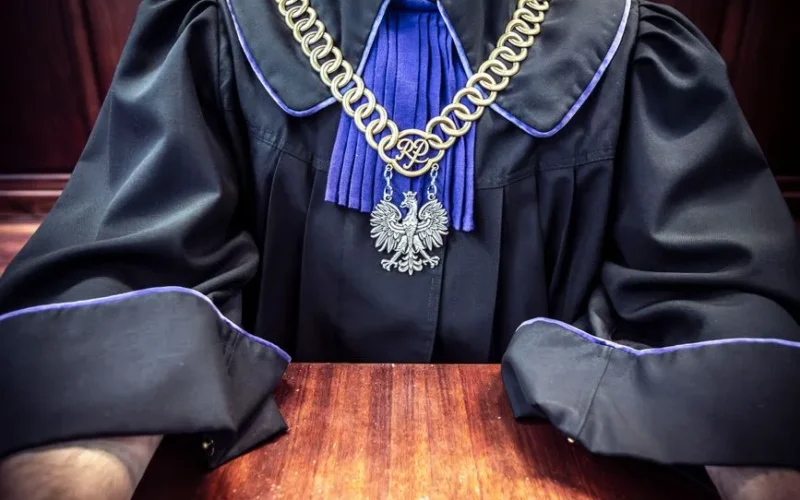 Close-up of a judge wearing a traditional robe with a golden chain medallion, seated at a courtroom table.