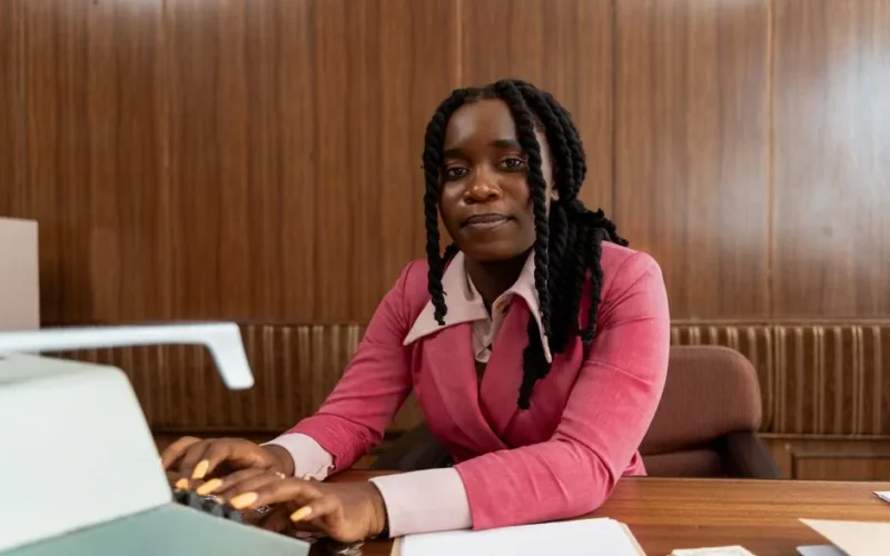 African American woman in a pink suit typing on a retro typewriter in an office setting.