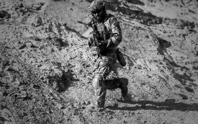 A soldier runs down a sandy hill in full combat gear under harsh desert sun.