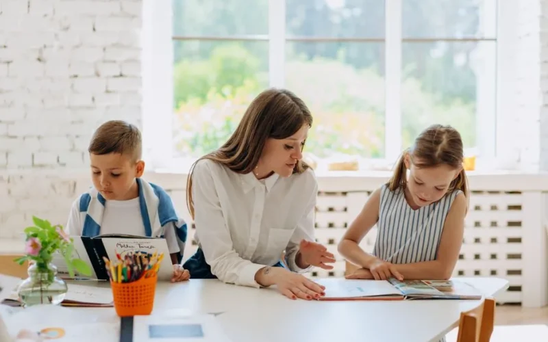 A female teacher helps two children learn by reading together in a bright classroom.