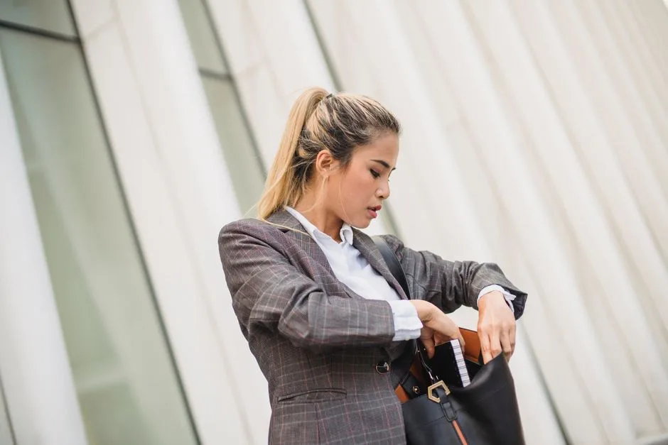 Confident young professional woman with ponytail and plaid blazer looking into her bag outdoors.
