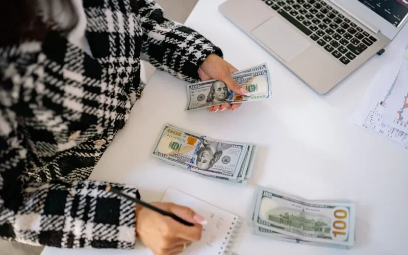A woman in a plaid coat counts American dollars at a desk with a notebook and laptop.