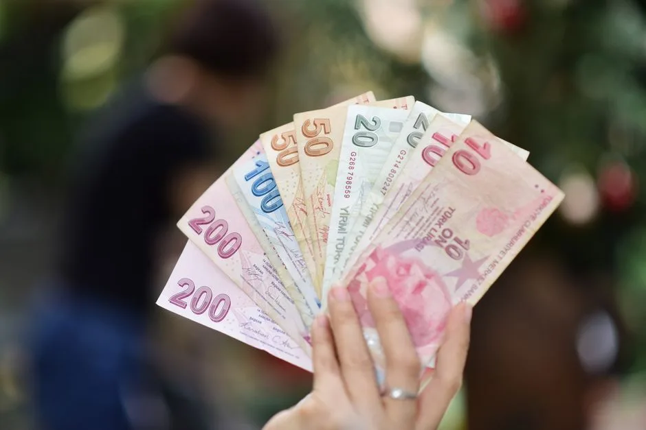 A hand holding various Turkish Lira banknotes with a blurred outdoor background.