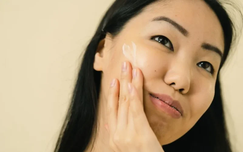 Woman with long black hair applying facial cream in studio. Focus on skincare and beauty routine.