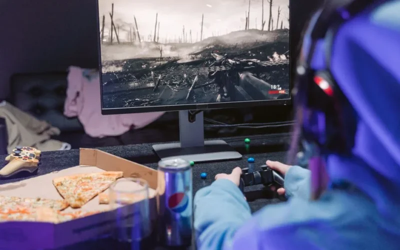 Teen playing video game with headset and drinks, indulging in pizza during gaming session.