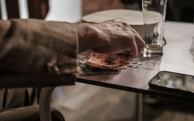 Hand counting euro banknotes on a table beside a phone and empty glass.