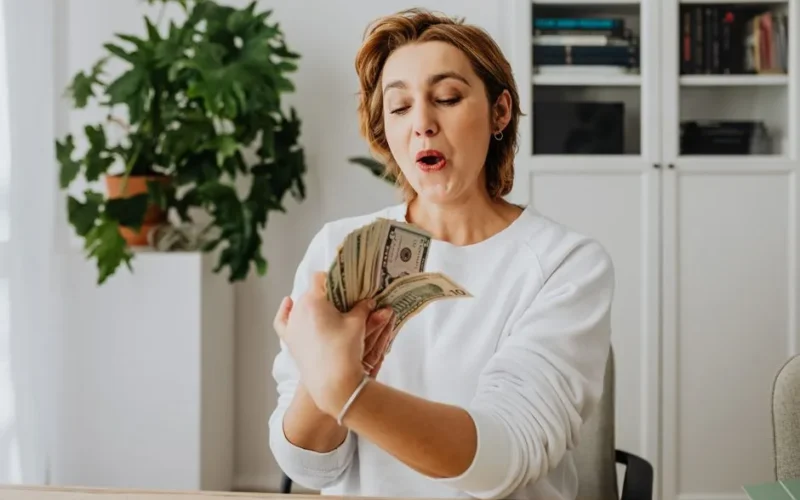 A woman in a white sweater excitedly counts American dollar bills indoors, reflecting joy and prosperity.