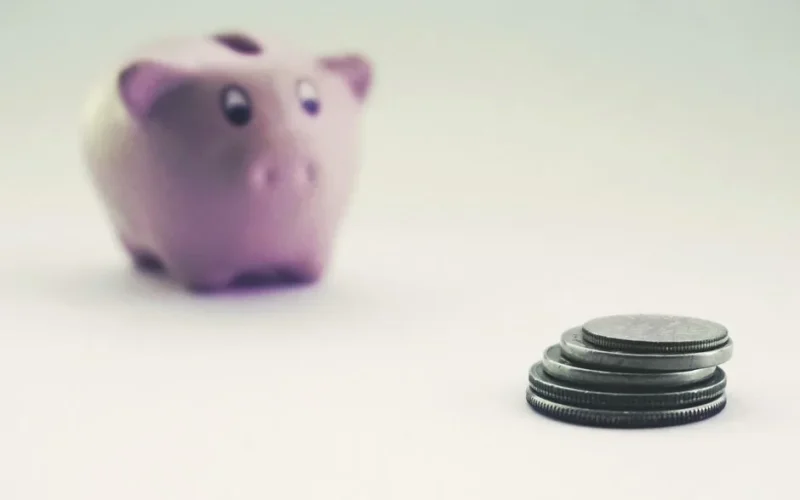 A pink piggy bank blurred in the background with stacked coins in the foreground on a white surface.