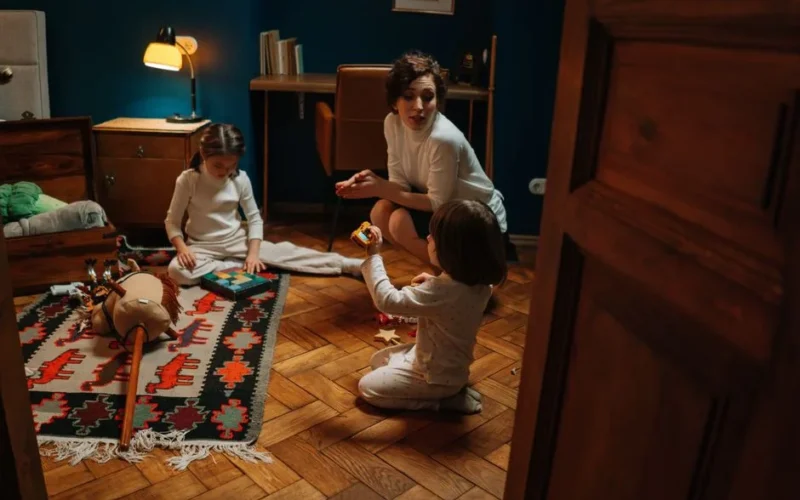 Woman and children playing with toys on a wooden floor in a warmly lit room.